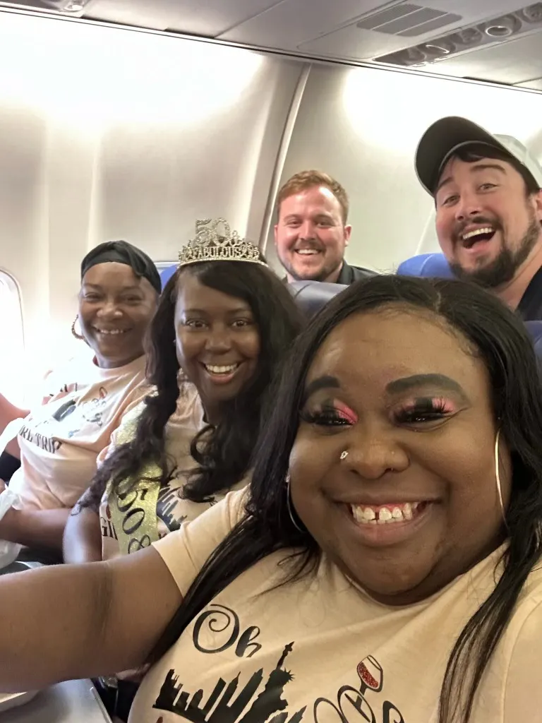 3 smiling black women on a plane headed to NYC for a birthday trip being photo bombed by 2 white men